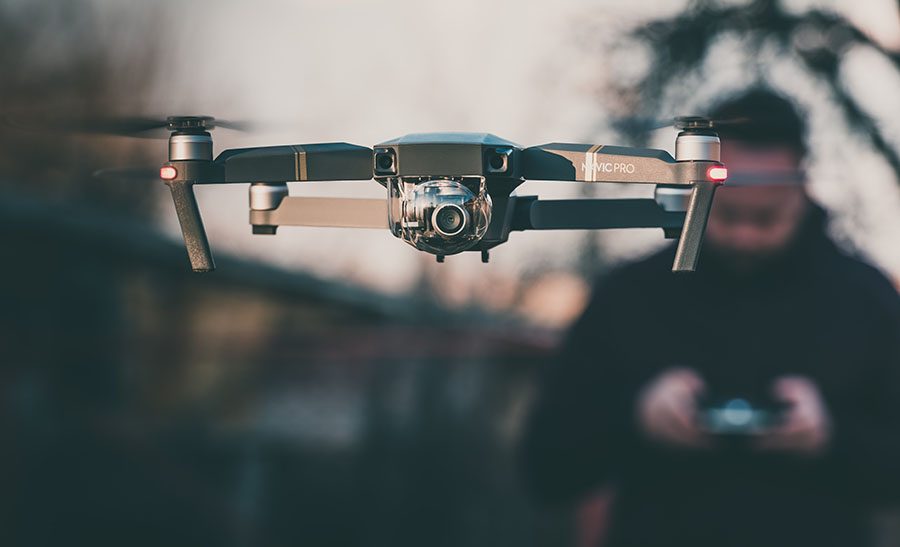 Roof Estimate. Image shows a drone in flight with a man out of focus behind it driving the drone.
