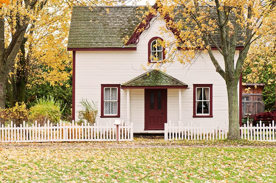 Roof estimate. Image shows a quaint house with white siding, red trim and a picket fence, with maple trees all around.