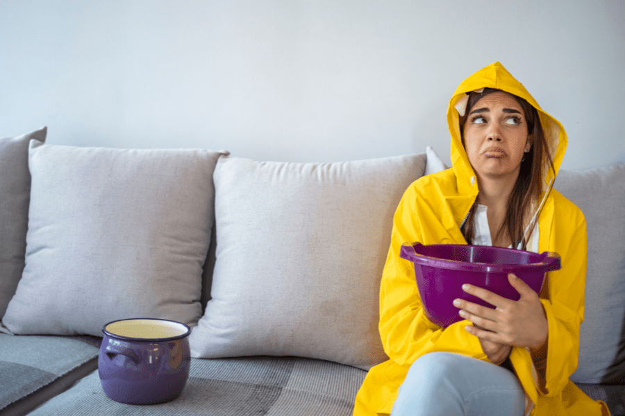 Roof leak showing a sad woman in a yellow raincoat sitting in her living room holding a purple bucket.